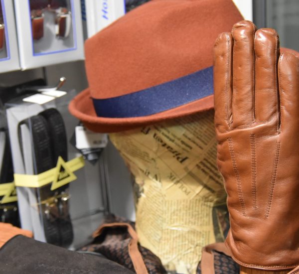 Ein brauner Lederhandschuh vor einem braunen Hut mit blauem Hutband. Im Hintergrund sind verpackte Hosenträger